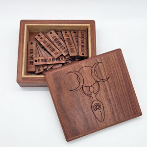 May include: A square wooden box with a lid featuring a laser-engraved triple moon goddess symbol. Inside the box are multiple small, rectangular wooden tags, each engraved with motivational text such as I Inspire People Daily.