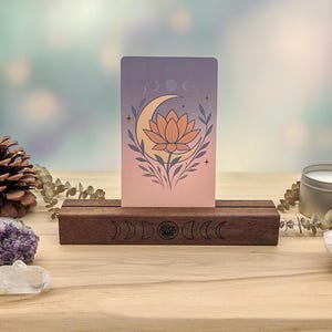 May include: A rectangular wooden card stand with a dark brown finish, featuring a laser-engraved moon phase and lotus design. A vertical card sits in the slot, displaying a gradient background with a central lotus flower and crescent moon.