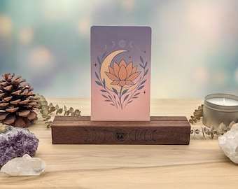 May include: A rectangular wooden card stand with a dark brown finish, featuring a laser-engraved moon phase and lotus design. A vertical card sits in the slot, displaying a gradient background with a central lotus flower and crescent moon.