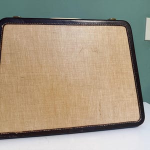May include: Vintage rectangular suitcase with a light tan woven fabric front and dark brown leather trim. The suitcase has a handle and metal hardware. The corners are rounded and the edges are stitched.