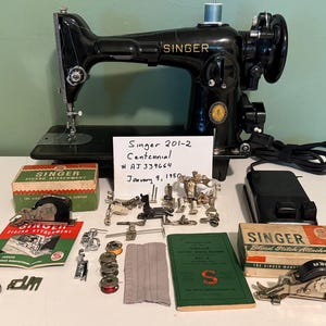 May include: A black Singer 201-2 Centennial sewing machine with a power cord, foot pedal, and various metal attachments. A white card reads Singer 201-2 Centennial # AJ 339664 January 9, 1950. Includes green boxes, instruction manuals, and bobbins.