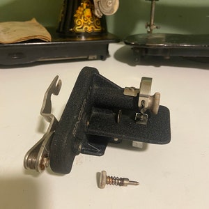 May include: A black Singer sewing machine foot with a metal spring and screw. The foot is made of metal and has a black finish. The spring is silver and the screw is silver with a black head.