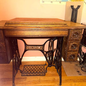 May include: A vintage Singer treadle sewing machine table featuring a dark oak wood top and a central drawer. The base is constructed from black cast iron with an intricate pedal mechanism. Three small drawers are located on each side of the unit.
