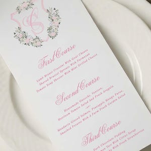 May include: A white menu card with pink text and floral details sits on white plates. The menu lists three courses: First, Second, and Third, with detailed descriptions of each dish. A floral crest with a pink initial is at the top.