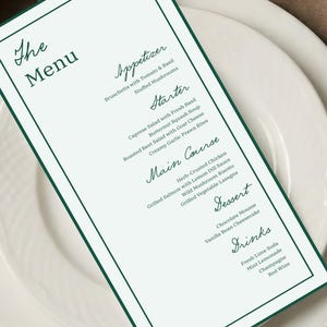 Puede incluir: Una tarjeta de menú con un borde verde y texto, colocada sobre platos blancos. El menú enumera aperitivos, entrantes, platos principales, postres y bebidas. El texto está en cursiva. El menú se titula "The Menu".