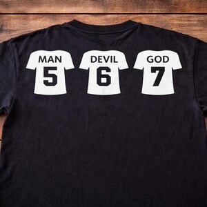 May include: Black t-shirt featuring three white jersey-style graphics. Each graphic displays a word and a number: "MAN 5", "DEVIL 6", and "GOD 7". The shirt is shown on a wooden surface.