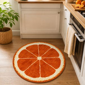 K&ouml;nnte beinhalten: Ein runder, orangefarbener und wei&szlig;er Teppich, der einer Zitrusfruchtscheibe &auml;hnelt. Der Teppich hat eine strukturierte Oberfl&auml;che und liegt auf einem Holzboden in einer K&uuml;che. Das Design des Teppichs zeigt eine leuchtend orangefarbene Mitte mit wei&szlig;en Segmenten und einer wei&szlig;en Schale.