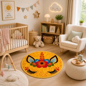 K&ouml;nnte beinhalten: Runder gelber Teppich mit einem Einhorn-Gesicht-Design. Er hat schwarze Wimpern, blaue Ohren und eine Blumenm&auml;hne in Rot, Orange und Blau. Der Teppich befindet sich in einem Kinderzimmer, zusammen mit einem Kinderbett, einem Schaukelstuhl und Holzregalen.