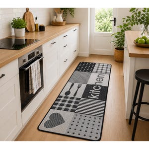 May include: A rectangular kitchen runner rug featuring a black and white patchwork design with patterns including stripes, checks, dots, and a heart shape. The word kitchen is printed in white text on a black background block on the rug.