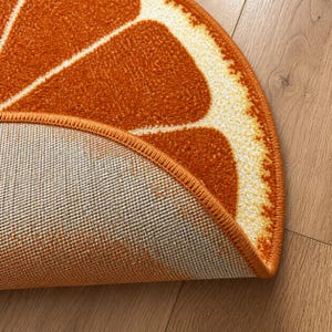 K&ouml;nnte beinhalten: Ein halbkreisf&ouml;rmiger Teppich mit einem Orangenscheiben-Design. Der Teppich hat eine leuchtend orange Farbe mit wei&szlig;en und gelben Akzenten, die die Segmente einer Orange nachahmen. Der Rand des Teppichs ist dunkler orange und die Unterseite ist beige.