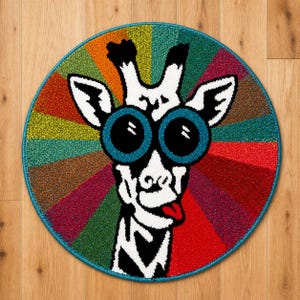 K&ouml;nnte beinhalten: Ein runder Teppich mit einem skurrilen Giraffen-Gesicht. Die Giraffe hat gro&szlig;e, blau umrandete Augen und eine rote Zunge, vor einem farbenfrohen, gestreiften Hintergrund. Der Teppich hat einen t&uuml;rkisfarbenen Rand. Die Giraffe hat schwarz-wei&szlig;e Markierungen.