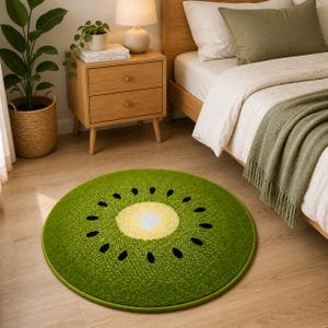 K&ouml;nnte beinhalten: Ein runder Teppich, der einer Kiwi-Frucht nachempfunden ist. Der Teppich hat einen gr&uuml;nen Au&szlig;enring, eine gelb-cremefarbene Mitte und schwarze Samen. Der Teppich liegt auf einem Holzboden, in der N&auml;he eines Bettes und eines Nachttisches.