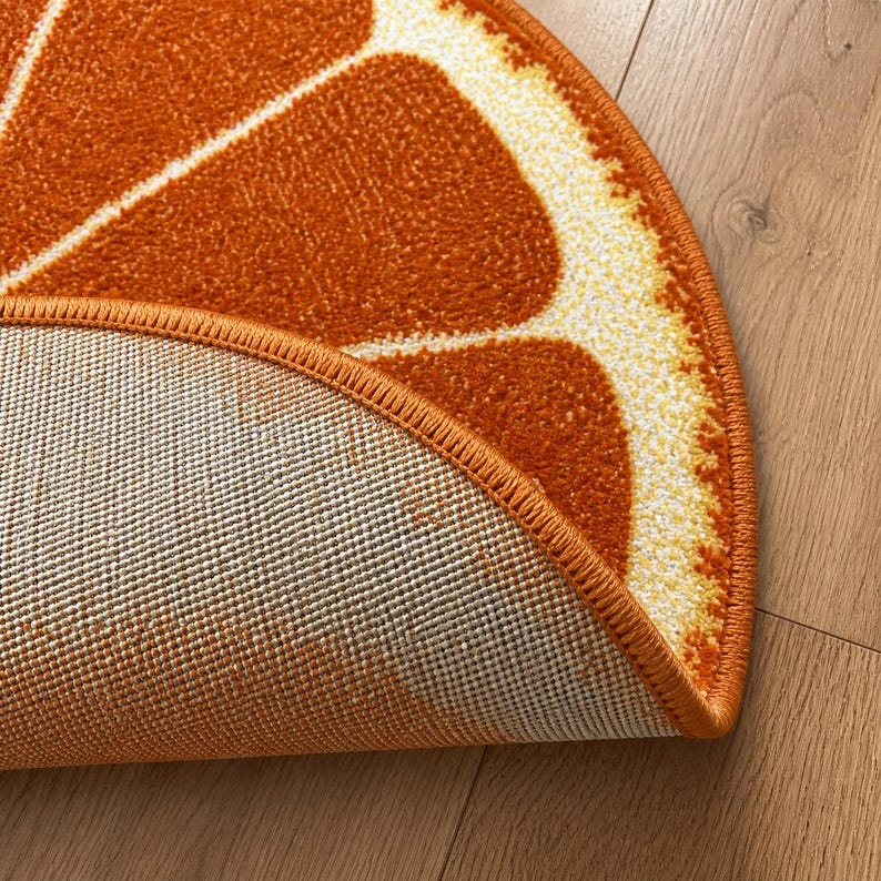 K&ouml;nnte beinhalten: Ein halbkreisf&ouml;rmiger Teppich mit einem Orangenscheiben-Design. Der Teppich hat eine leuchtend orange Farbe mit wei&szlig;en und gelben Akzenten, die die Segmente einer Orange nachahmen. Der Rand des Teppichs ist dunkler orange und die Unterseite ist beige.