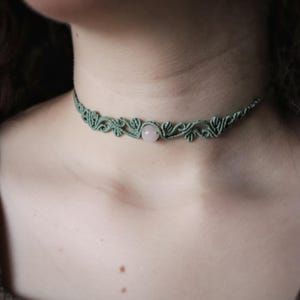 May include: A green macrame choker necklace with a central rose quartz stone. The necklace has detailed knotwork and is designed to sit around the neck. The colour is a muted green.