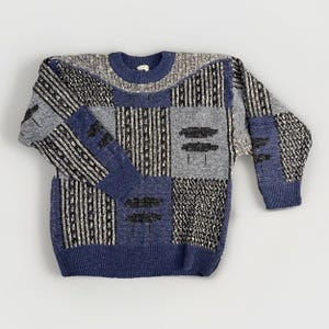 May include: A long-sleeve crew neck sweater featuring a patchwork design of blue, black, and grey knit panels. The garment displays alternating textures including vertical stripes, solid blue blocks, and speckled patterns with a ribbed collar and cuffs.