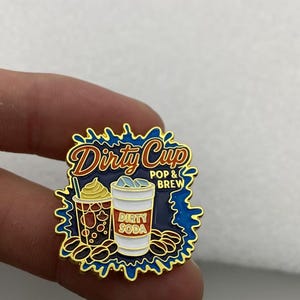 May include: A gold-rimmed enamel pin featuring a dark blue splash background with the text Dirty Cup POP & BREW. The design includes a brown coffee cup with whipped cream and a white soda cup labeled DIRTY SODA, surrounded by scattered coffee beans.
