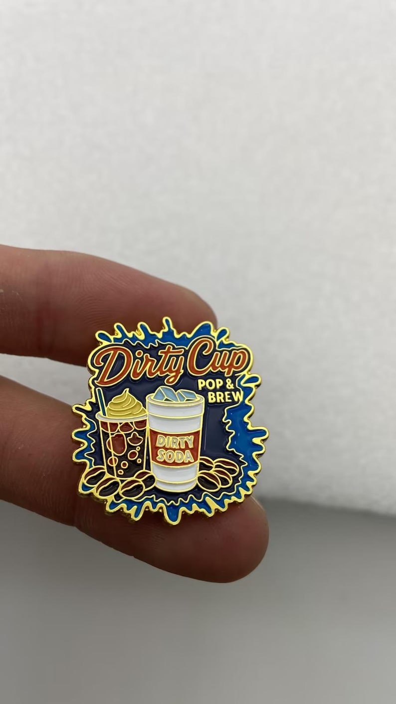 May include: A gold-rimmed enamel pin featuring a dark blue splash background with the text Dirty Cup POP & BREW. The design includes a brown coffee cup with whipped cream and a white soda cup labeled DIRTY SODA, surrounded by scattered coffee beans.