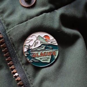 May include: A round enamel pin featuring a mountain landscape with the text "GLACIER NATIONAL PARK". The pin has a white, green, orange, and teal color scheme, with a gold-colored border. The pin is attached to a green jacket.