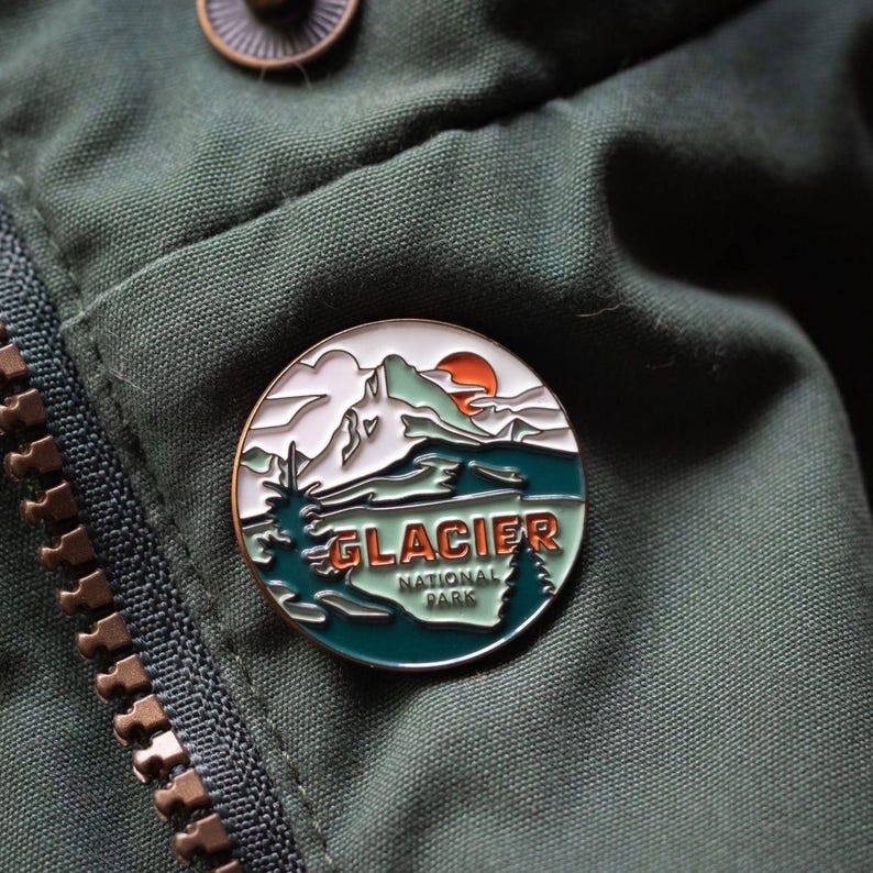 May include: A round enamel pin featuring a mountain landscape with the text "GLACIER NATIONAL PARK". The pin has a white, green, orange, and teal color scheme, with a gold-colored border. The pin is attached to a green jacket.