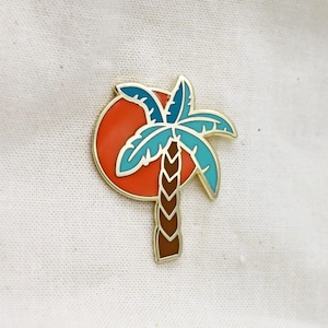 May include: A gold-toned enamel pin featuring a palm tree silhouette against a red sun. The palm tree has blue and teal fronds and a brown trunk. The pin is set against a white fabric background.