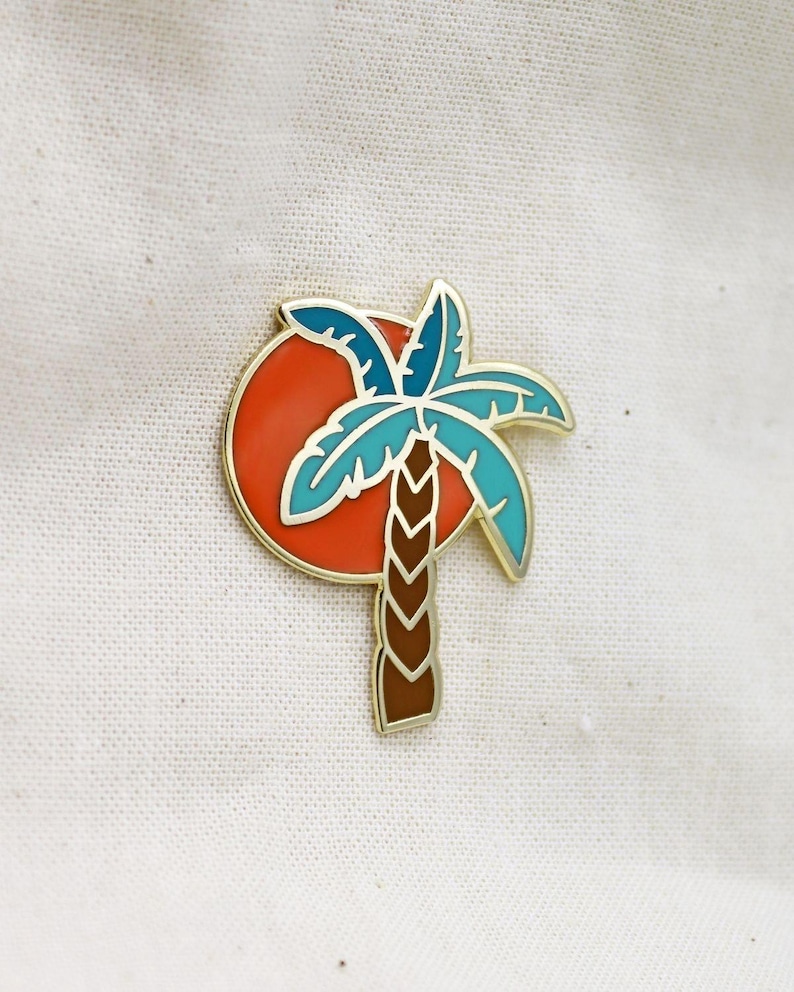 May include: A gold-toned enamel pin featuring a palm tree silhouette against a red sun. The palm tree has blue and teal fronds and a brown trunk. The pin is set against a white fabric background.