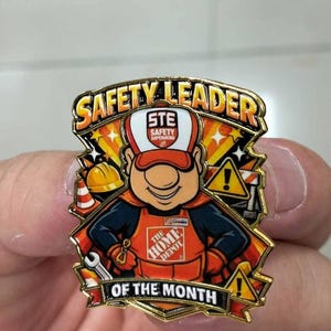 May include: A gold-toned metal enamel pin featuring a cartoon character wearing a Home Depot apron and cap. The text reads SAFETY LEADER OF THE MONTH. The design includes a yellow hard hat, a traffic cone, a wrench, and two yellow warning signs.