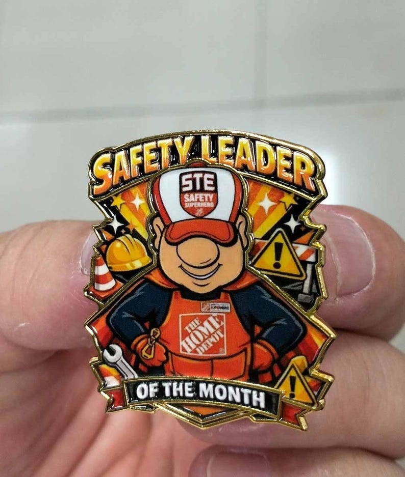 May include: A gold-toned metal enamel pin featuring a cartoon character wearing a Home Depot apron and cap. The text reads SAFETY LEADER OF THE MONTH. The design includes a yellow hard hat, a traffic cone, a wrench, and two yellow warning signs.