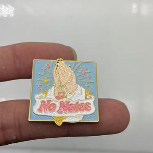 May include: A square enamel pin with a light blue background featuring a pair of hands in a praying position. The hands are adorned with a beaded necklace and a pink musical note charm. A white banner with pink glitter text reads No Notes.