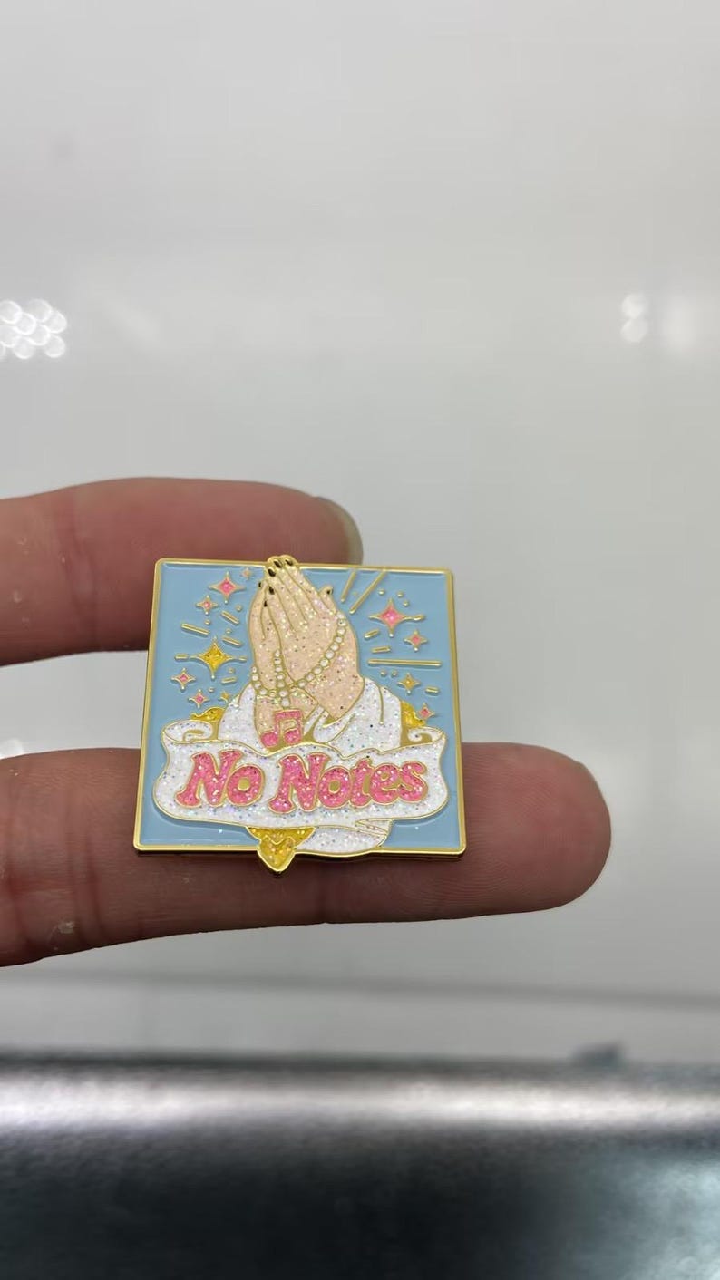 May include: A square enamel pin with a light blue background featuring a pair of hands in a praying position. The hands are adorned with a beaded necklace and a pink musical note charm. A white banner with pink glitter text reads No Notes.