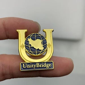 May include: A gold-toned metal lapel pin shaped like the letter U, featuring a dark blue globe with a gold map silhouette in the center. The pin includes a rectangular base with the text UnityBridge in white lettering against a dark blue background.