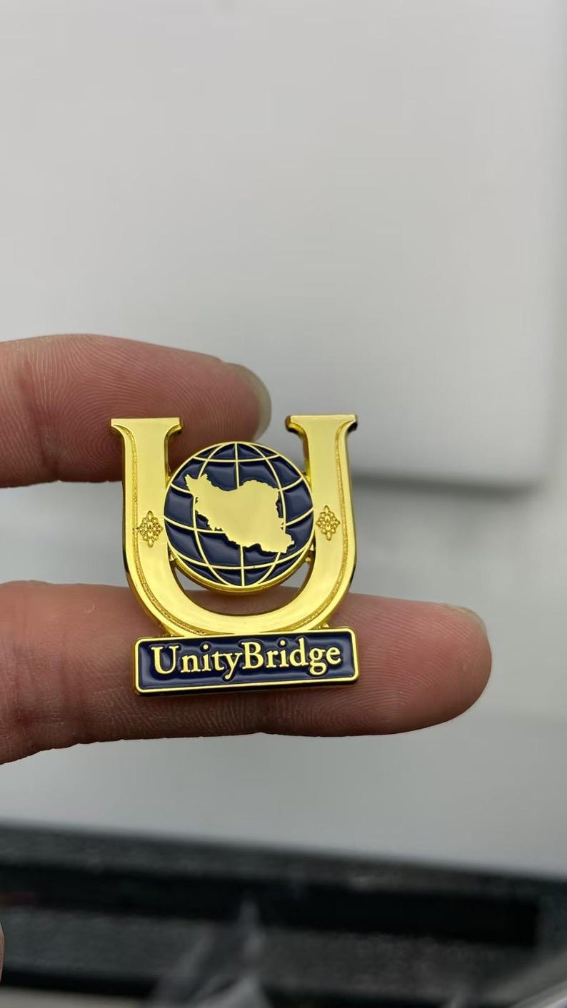 May include: A gold-toned metal lapel pin shaped like the letter U, featuring a dark blue globe with a gold map silhouette in the center. The pin includes a rectangular base with the text UnityBridge in white lettering against a dark blue background.