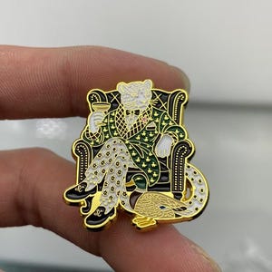 May include: A gold-toned metal enamel pin featuring a white leopard wearing a green patterned smoking jacket and white star-patterned trousers, sitting in a black armchair while holding a glass, with a mallard duck resting at its feet.