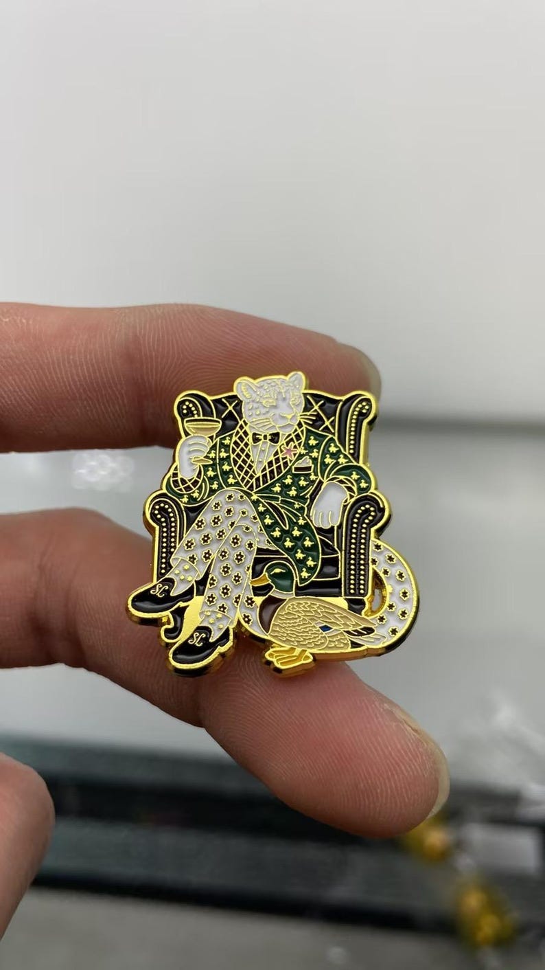 May include: A gold-toned metal enamel pin featuring a white leopard wearing a green patterned smoking jacket and white star-patterned trousers, sitting in a black armchair while holding a glass, with a mallard duck resting at its feet.