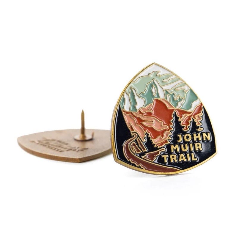 May include: A John Muir Trail enamel pin with a mountain and tree design. The pin is triangular with a gold border, featuring red, green, and black colors. The text "JOHN MUIR TRAIL" is displayed on the pin.