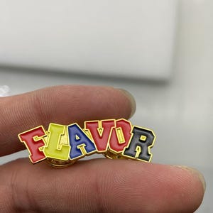 May include: A gold-toned metal enamel pin featuring the word FLAVOR in block lettering. Each letter is filled with a distinct color: F is red, L is yellow, A is blue, V is red, O is yellow, and R is black. The pin has a polished gold metal border.