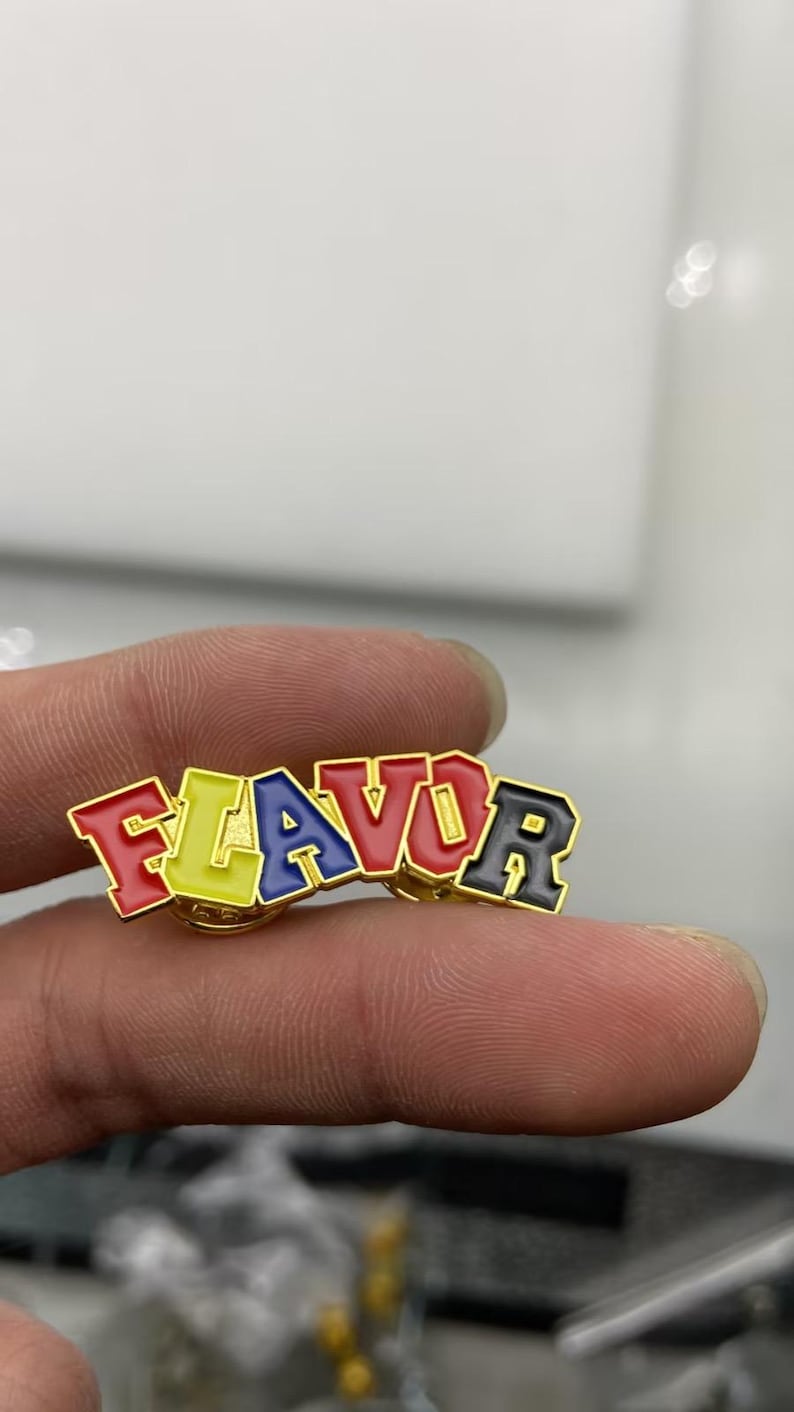May include: A gold-toned metal enamel pin featuring the word FLAVOR in block lettering. Each letter is filled with a distinct color: F is red, L is yellow, A is blue, V is red, O is yellow, and R is black. The pin has a polished gold metal border.