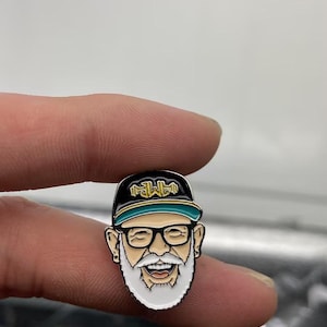 May include: A metal enamel pin featuring a cartoon face with a white beard, black-rimmed glasses, and a black baseball cap with a teal brim. The cap displays the gold text "AMP" in a stylized font. The character has a wide, smiling expression.