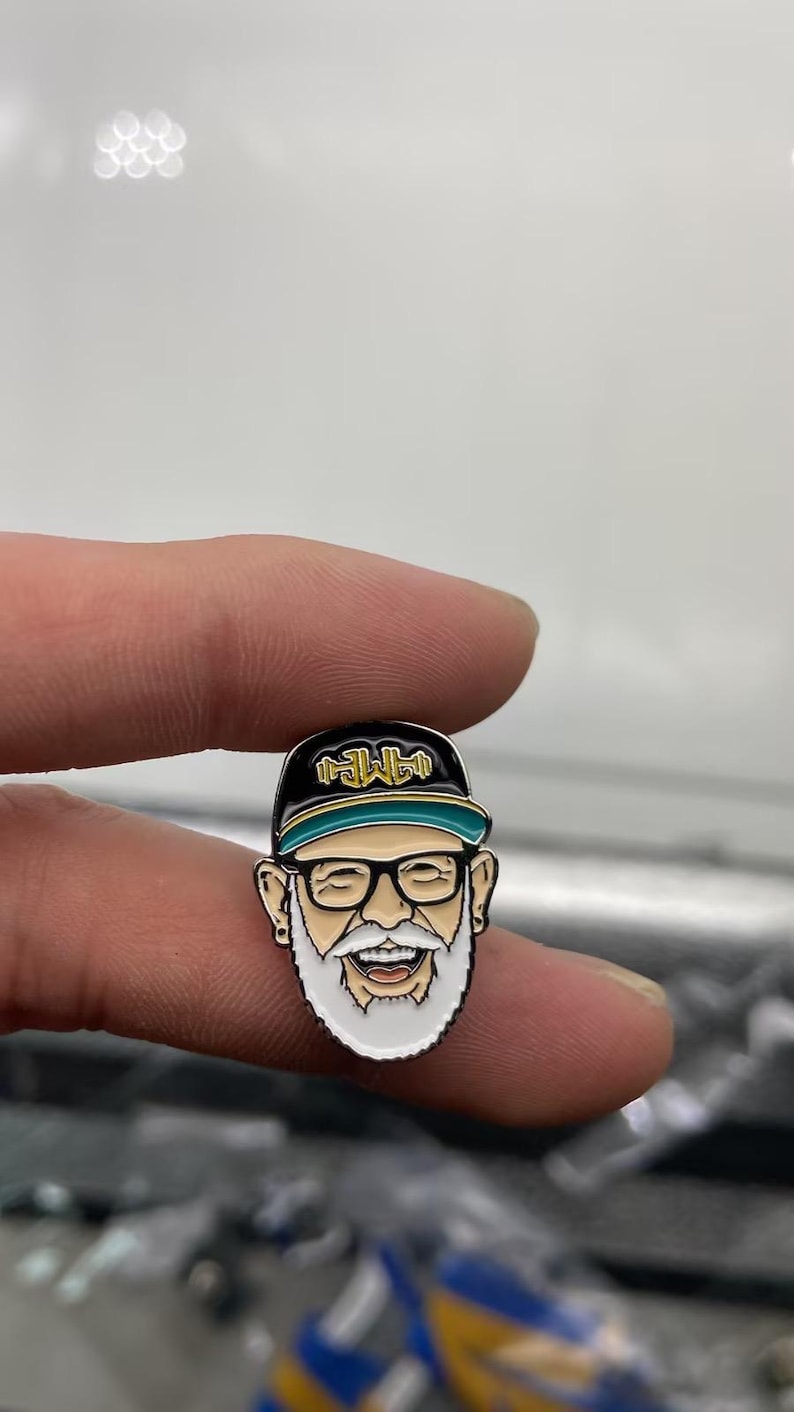 May include: A metal enamel pin featuring a cartoon face with a white beard, black-rimmed glasses, and a black baseball cap with a teal brim. The cap displays the gold text "AMP" in a stylized font. The character has a wide, smiling expression.