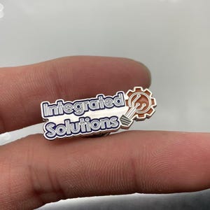 May include: A metal lapel pin featuring the text Integrated Solutions in blue and white lettering. To the right of the text is a copper-colored gear icon containing a white lightbulb graphic with circuit board lines extending from the base.