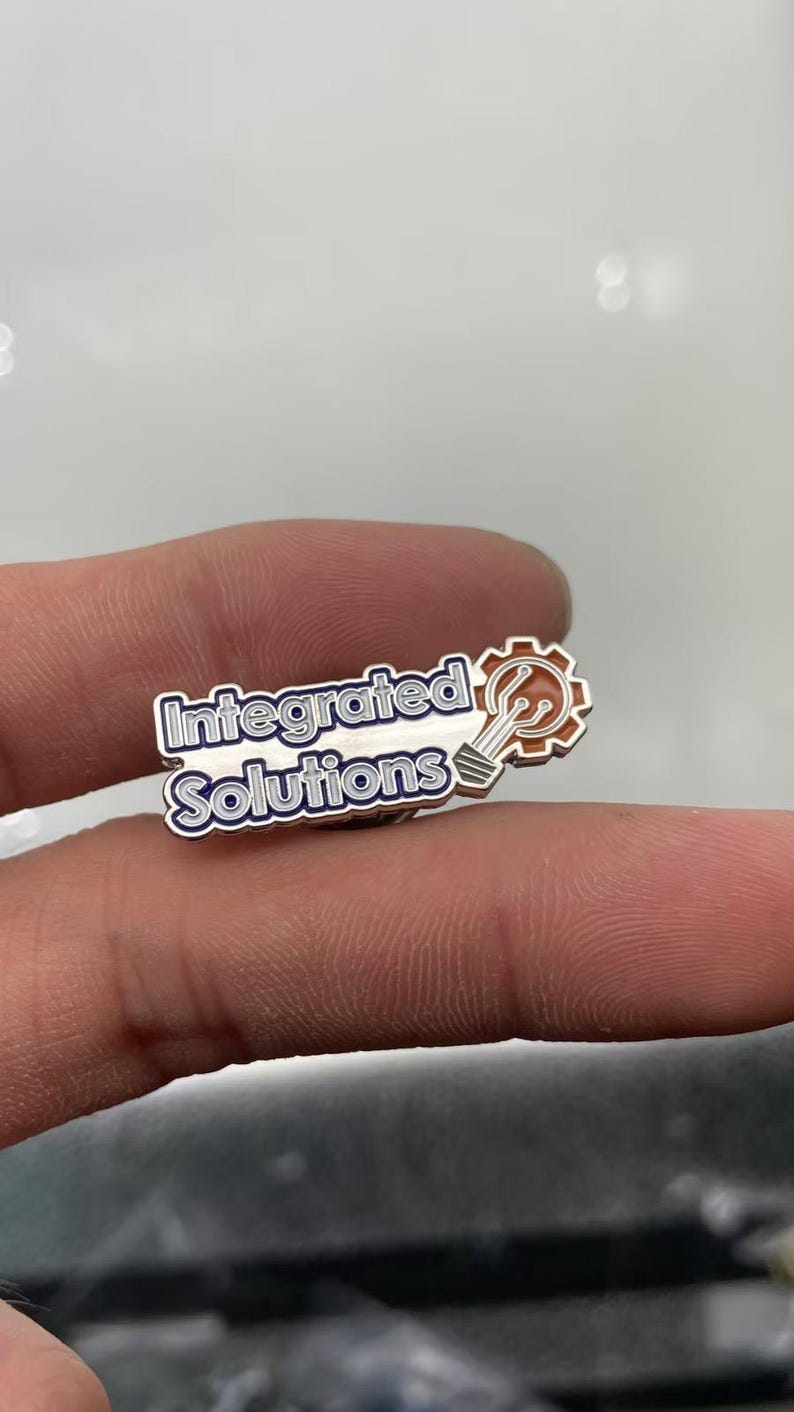 May include: A metal lapel pin featuring the text Integrated Solutions in blue and white lettering. To the right of the text is a copper-colored gear icon containing a white lightbulb graphic with circuit board lines extending from the base.