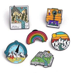 May include: A collection of enamel pins with outdoor and travel themes. Designs include a mountain landscape, a rainbow, a compass, a camera, and a mountain scene inside a book. Each pin features vibrant colors and detailed artwork.