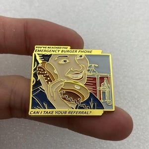 May include: A square enamel pin featuring a person holding a burger to their ear like a telephone. The text reads YOU'VE REACHED THE EMERGENCY BURGER PHONE and CAN I TAKE YOUR REFERRAL? The design uses gold, tan, dark blue, red, and grey colors.