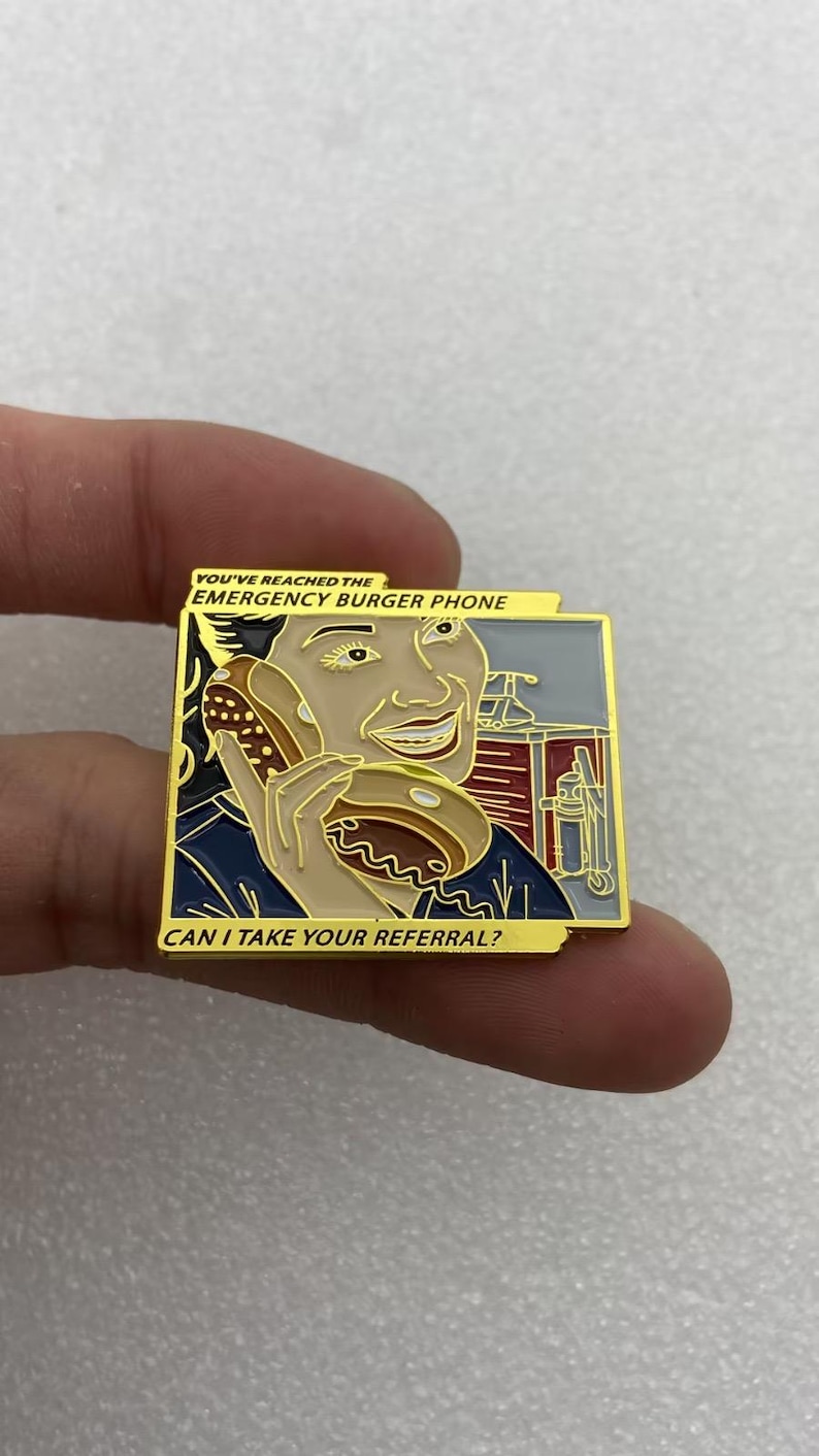 May include: A square enamel pin featuring a person holding a burger to their ear like a telephone. The text reads YOU'VE REACHED THE EMERGENCY BURGER PHONE and CAN I TAKE YOUR REFERRAL? The design uses gold, tan, dark blue, red, and grey colors.