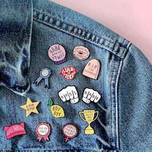 May include: A collection of enamel pins with various designs and text, including phrases like "Babe with the Power," "Boss," and "Babes Against Bigots." The pins are attached to a denim jacket, showcasing a variety of colors and shapes.