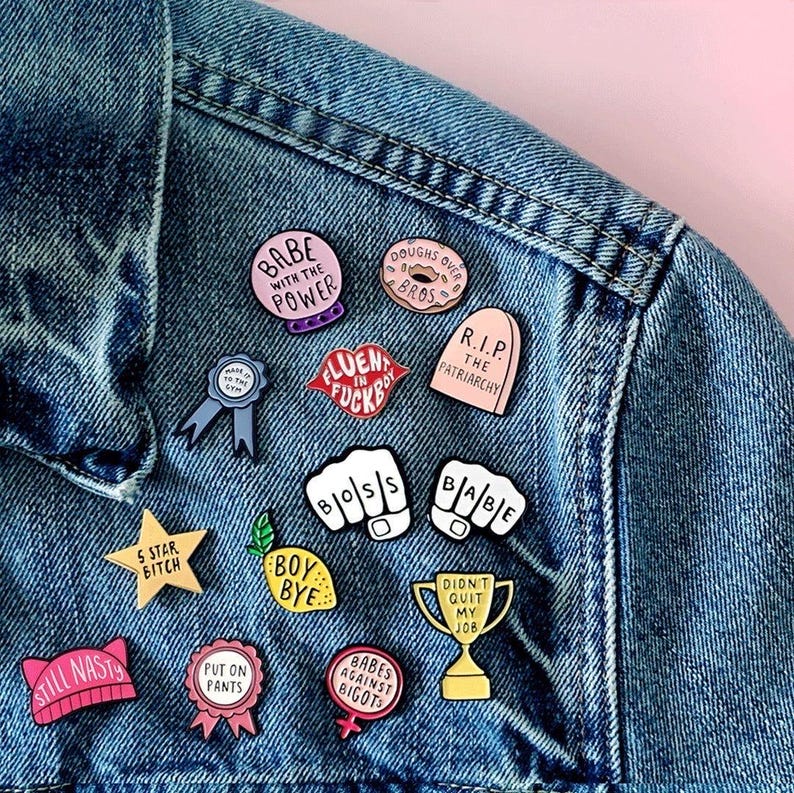 May include: A collection of enamel pins with various designs and text, including phrases like "Babe with the Power," "Boss," and "Babes Against Bigots." The pins are attached to a denim jacket, showcasing a variety of colors and shapes.