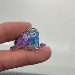 May include: A metal enamel pin featuring two owls perched on a branch. One owl is purple and the other is blue, both with closed eyes. The branch is brown with small pink flowers attached to it. The pin has a silver-toned metal outline.