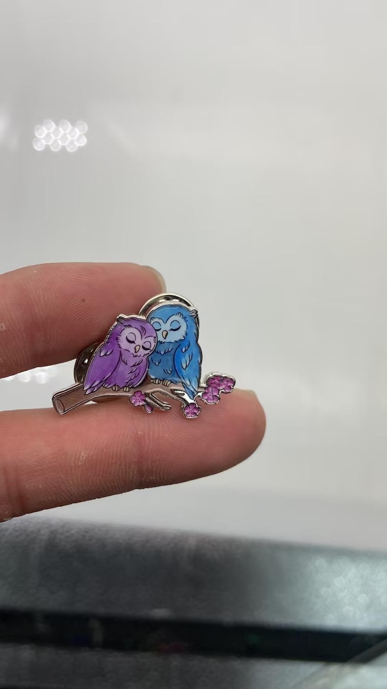 May include: A metal enamel pin featuring two owls perched on a branch. One owl is purple and the other is blue, both with closed eyes. The branch is brown with small pink flowers attached to it. The pin has a silver-toned metal outline.