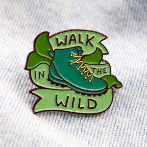 May include: A green enamel pin featuring a hiking boot with yellow laces, set against a green banner that reads "WALK IN THE WILD." The pin has a dark brown outline and green leaves. The pin is on a light blue denim background.