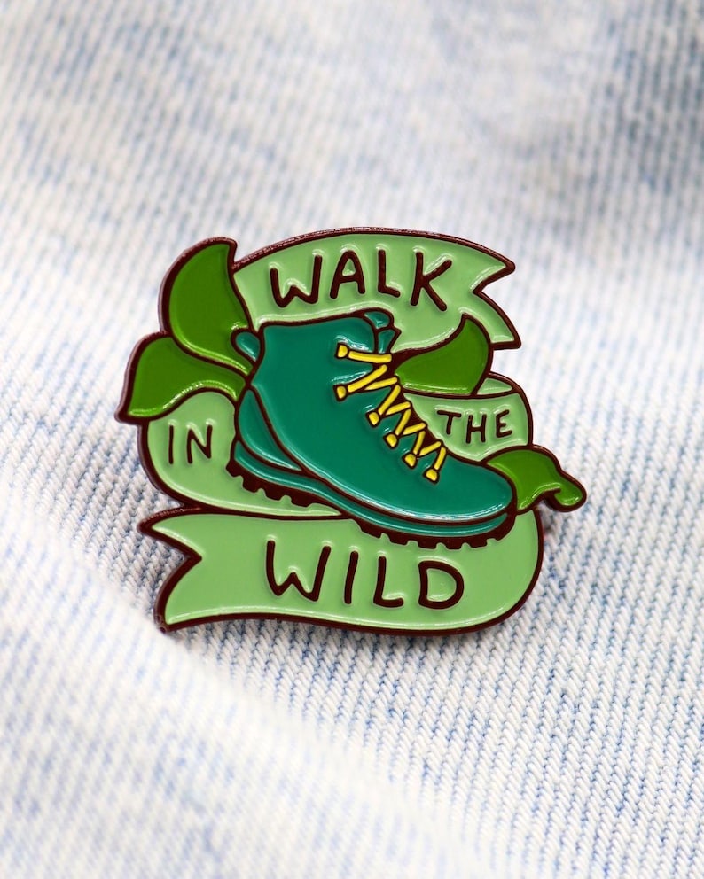 May include: A green enamel pin featuring a hiking boot with yellow laces, set against a green banner that reads "WALK IN THE WILD." The pin has a dark brown outline and green leaves. The pin is on a light blue denim background.