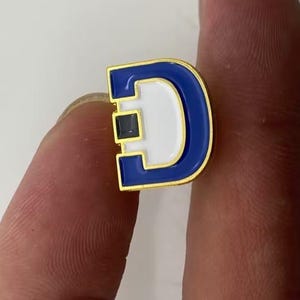 May include: A gold-toned metal lapel pin shaped like the letter D, featuring a bold blue enamel border and a white enamel center. A small, square, dark blue accent is embedded on the left side of the letter, adding a distinct detail to the design.